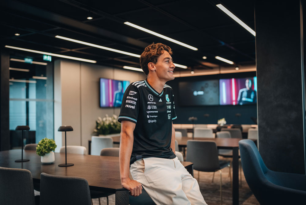 George Russell, piloto del equipo Mercedes-AMG PETRONAS Formula One Team, está sentado de manera relajada sobre una mesa en un espacio moderno y elegante del equipo, ya sea una oficina o un área de hospitalidad. Sonríe y lleva puesta la camisa oficial del equipo con los logotipos de los sponsors visibles, como Einhell, IWC y CrowdStrike. En las pantallas del fondo se puede ver su imagen.