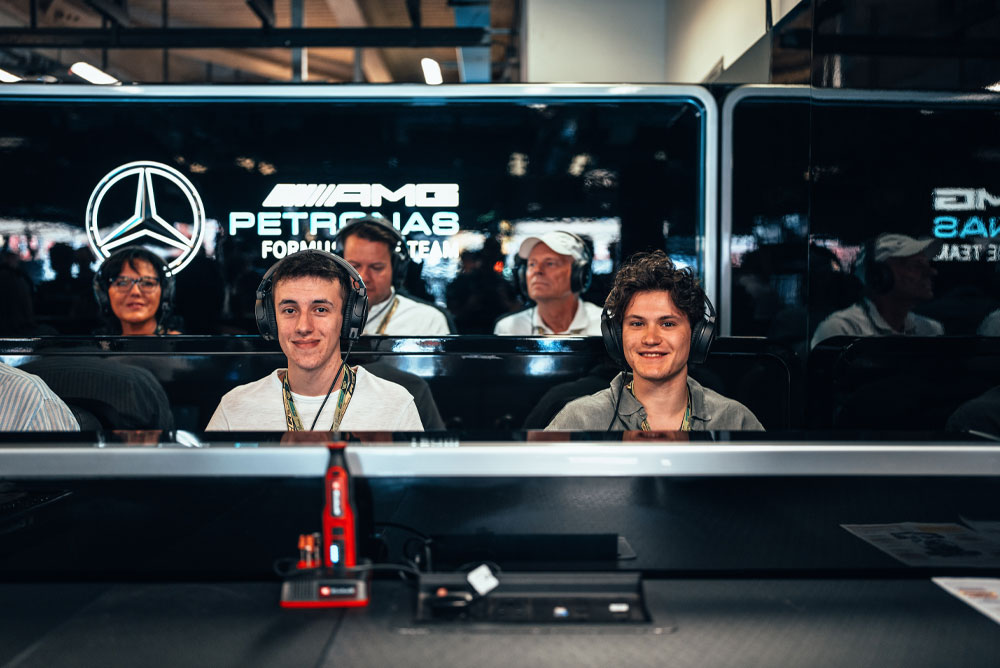 Dos hombres están sentados en el taller del equipo Mercedes-AMG PETRONAS Formula One, usando audífonos y disfrutando la experiencia detrás de escena. En primer plano se aprecia un dispositivo rojo de Einhell.
