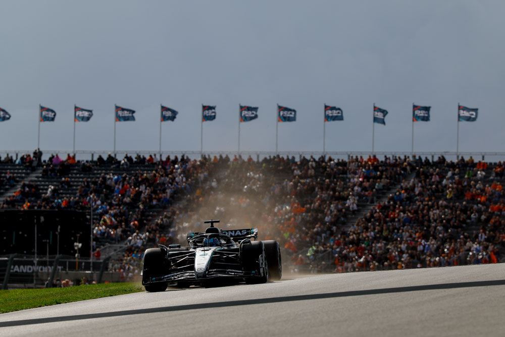 Un auto del equipo Mercedes-AMG PETRONAS Formula One pasa frente a tribunas repletas durante una carrera. Los fanáticos agitan banderas al fondo, creando una atmósfera llena de energía.