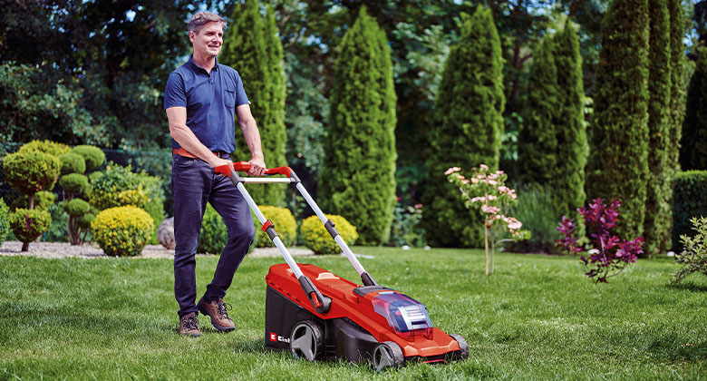 A man mowing the green lawn in a garden with a red and black cordless lawn mower.
