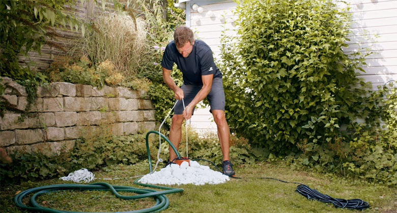 A man standing bent over a pipe in the garden, a rope in his hand to which a deep well pump is attached.