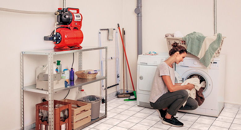 A woman loading a washing machine, next to her is a house water pump on a shelf.