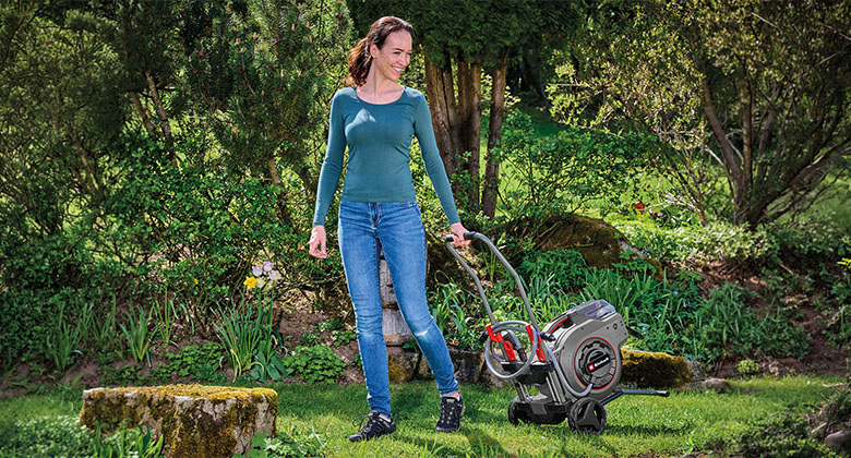 Frau schiebt einen kompakten Rasenmäher mit schwarzem und rotem Gehäuse über eine grüne Rasenfläche im Garten.