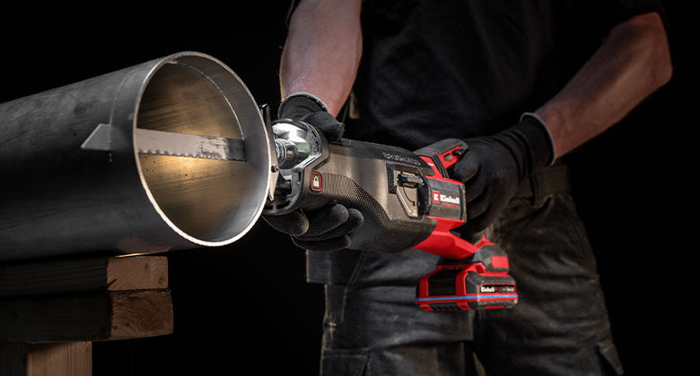 Person mit Handschuhen schneidet ein Metallrohr mit einer roten Akku-Säbelsäge auf einer Holzauflage.